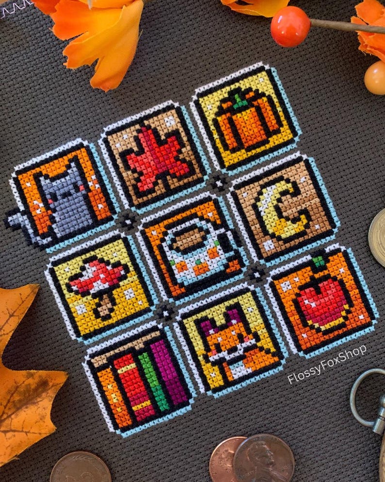May include: A cross-stitch piece with nine square panels depicting autumn symbols. The panels show a cat, maple leaf, pumpkin, crescent moon, mushroom, coffee cup, apple, books, and a fox. The piece is set against a grey background.