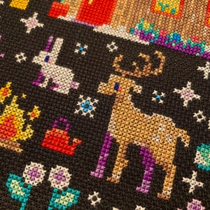May include: A black cross-stitch design featuring a white rabbit, a brown reindeer, and various flowers and plants. The background is black with white stars.