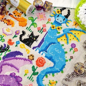 May include: A detailed cross-stitch design featuring a blue dragon with yellow accents, surrounded by flowers and small animal figures. A gold thread spool and dice are also visible, suggesting a crafting or gaming theme.