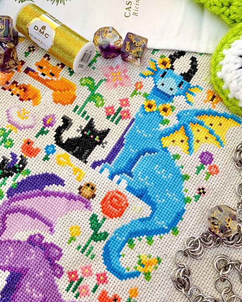 May include: A detailed cross-stitch design featuring a blue dragon with yellow accents, surrounded by flowers and small animal figures. A gold thread spool and dice are also visible, suggesting a crafting or gaming theme.