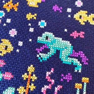May include: A close-up of a blue fabric with colorful cross-stitch embroidery. The design features a green frog, yellow and orange fish, and pink and purple flowers.