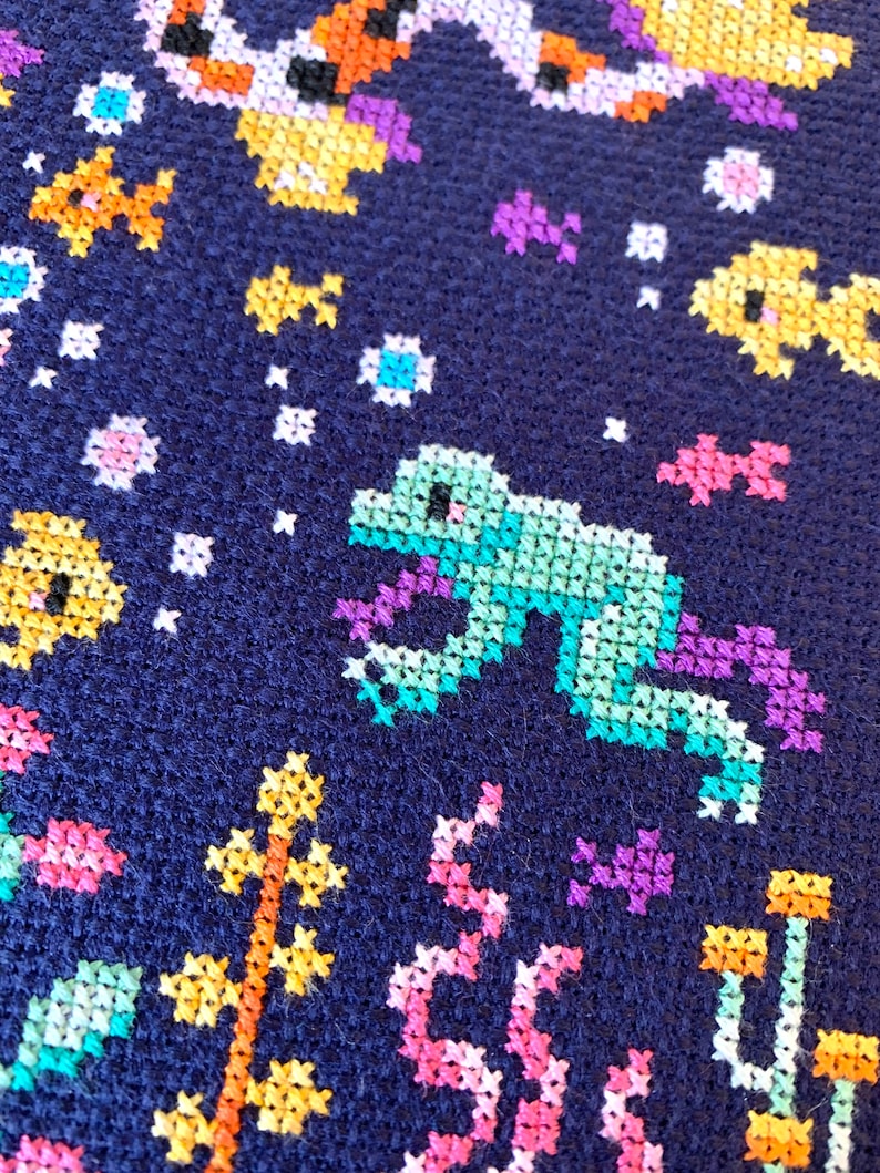 May include: A close-up of a blue fabric with colorful cross-stitch embroidery. The design features a green frog, yellow and orange fish, and pink and purple flowers.