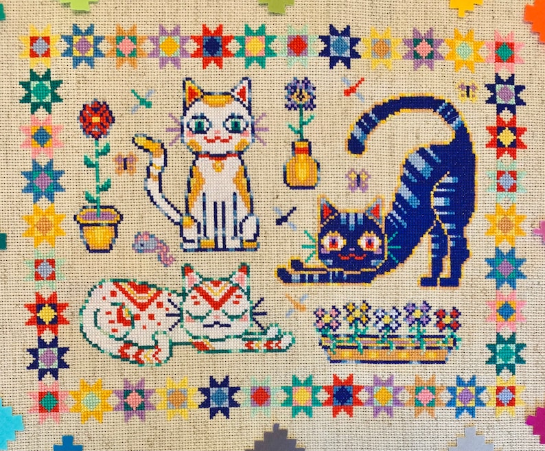May include: A cross-stitch embroidery featuring three cats. The cats are a white and yellow cat with a red collar, a blue and white cat with a yellow collar, and a white and red cat with a green collar. The cats are surrounded by a colorful border of stars and flowers.