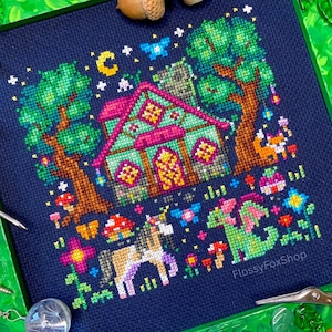 May include: A cross-stitch design depicting a fantasy cottage scene with a unicorn and dragon. The artwork is framed in green and includes trees, mushrooms, and a night sky with a moon and stars. The text "FlossyFoxShop" is visible.