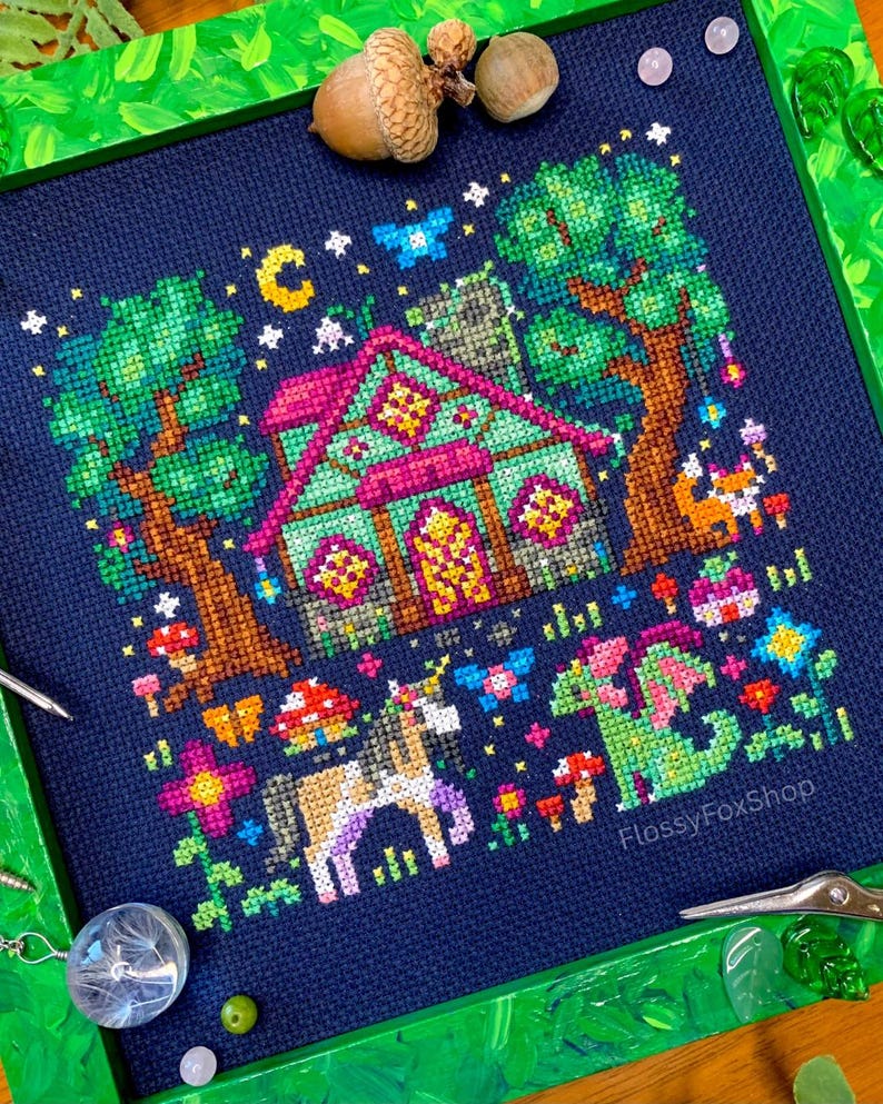 May include: A cross-stitch design depicting a fantasy cottage scene with a unicorn and dragon. The artwork is framed in green and includes trees, mushrooms, and a night sky with a moon and stars. The text "FlossyFoxShop" is visible.