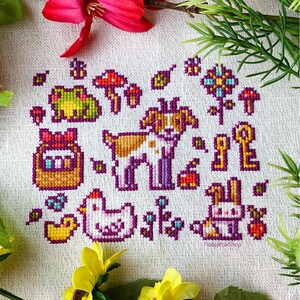 K&ouml;nnte beinhalten: Kreuzstichmuster mit verschiedenen Tieren und Objekten, darunter ein Hund, ein Kaninchen, ein Frosch, ein Huhn, ein Korb mit Eiern, Pilze, Schl&uuml;ssel und Blumen. Das Muster ist auf wei&szlig;em Aida-Stoff mit bunten F&auml;den gestickt.