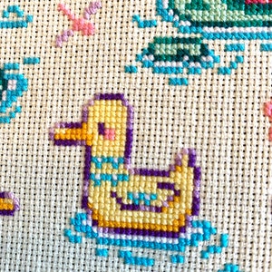 May include: A yellow duck with a purple and blue border is embroidered on a white fabric. The duck is swimming in blue water.