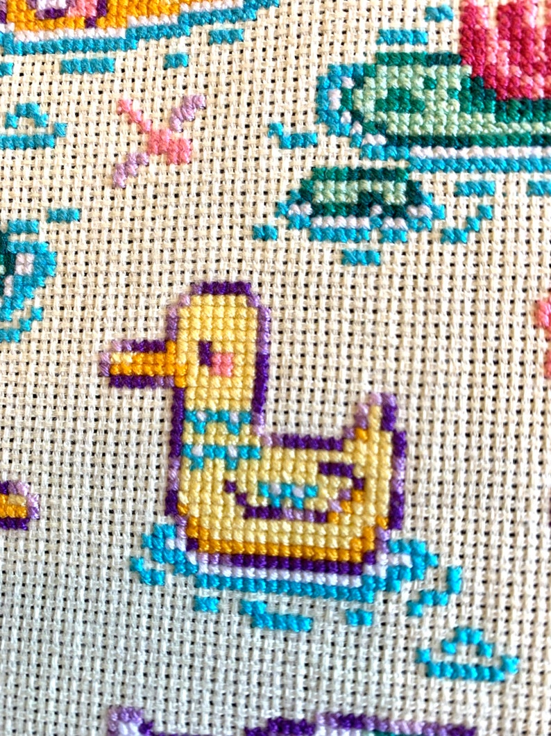 May include: A yellow duck with a purple and blue border is embroidered on a white fabric. The duck is swimming in blue water.