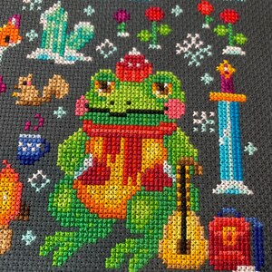 May include: A green frog wearing a red scarf and a red hat is holding a yellow guitar. The frog is sitting on a gray background with snowflakes and other winter-themed designs.