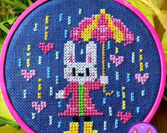 May include: A cross-stitch embroidery hoop with a blue background featuring a white bunny wearing a pink and green coat and holding a pink and yellow umbrella. The bunny is surrounded by pink hearts and blue raindrops.
