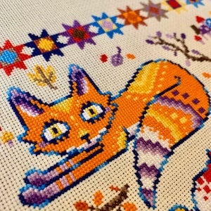 Puede incluir: Un patr&oacute;n de punto de cruz con un gato naranja de dibujos animados con rayas moradas y azules. El gato est&aacute; acostado de lado con la cabeza girada hacia la izquierda. El fondo es una tela de color crema con un borde de estrellas de colores.