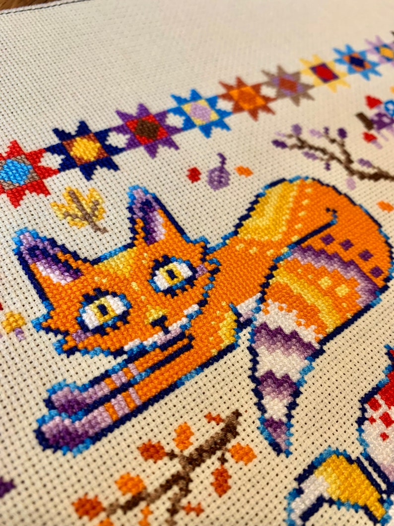 Puede incluir: Un patr&oacute;n de punto de cruz con un gato naranja de dibujos animados con rayas moradas y azules. El gato est&aacute; acostado de lado con la cabeza girada hacia la izquierda. El fondo es una tela de color crema con un borde de estrellas de colores.