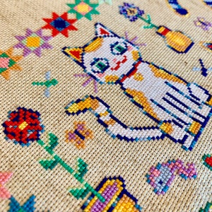 May include: Cross-stitch embroidery featuring a white cat with orange markings, sitting with its tail curled around its body. The cat is surrounded by colorful flowers and other designs.