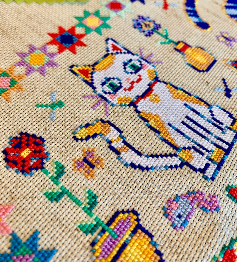 May include: Cross-stitch embroidery featuring a white cat with orange markings, sitting with its tail curled around its body. The cat is surrounded by colorful flowers and other designs.