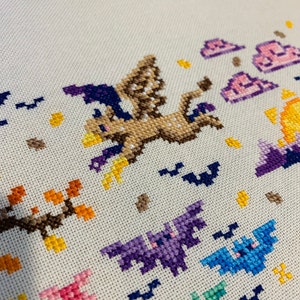 May include: Cross-stitch embroidery featuring a brown and purple flying creature with a witch's hat, surrounded by bats and clouds. The background is a light beige fabric.