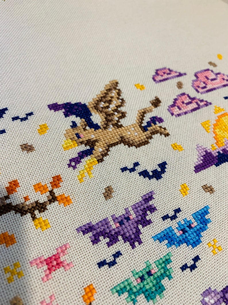 May include: Cross-stitch embroidery featuring a brown and purple flying creature with a witch's hat, surrounded by bats and clouds. The background is a light beige fabric.