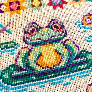 May include: A cross-stitch pattern featuring a green frog with yellow eyes and a yellow belly sitting on a green lily pad. The frog is surrounded by colorful flowers and stars.