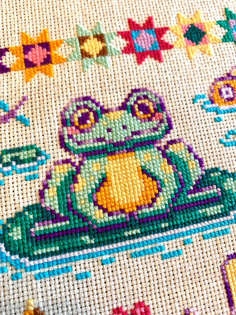 May include: A cross-stitch pattern featuring a green frog with yellow eyes and a yellow belly sitting on a green lily pad. The frog is surrounded by colorful flowers and stars.