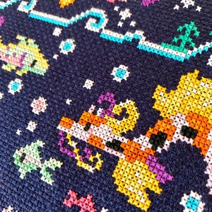 May include: Close-up of a colorful cross-stitch pattern on a dark blue fabric. The pattern features a giraffe, a fish, and a palm tree, all in a pixelated style.