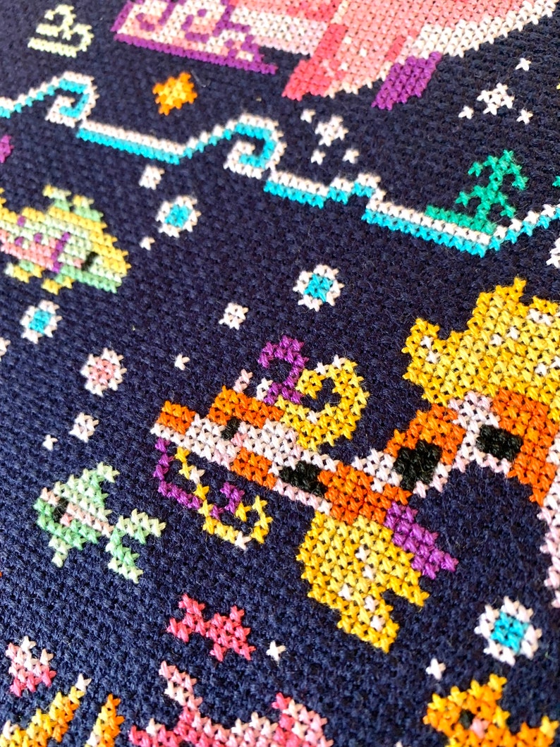 May include: Close-up of a colorful cross-stitch pattern on a dark blue fabric. The pattern features a giraffe, a fish, and a palm tree, all in a pixelated style.
