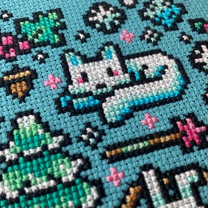 Pu&ograve; includere: Motivo a punto croce su tessuto azzurro chiaro raffigurante un unicorno bianco con accenti blu, circondato da stelle e altri elementi decorativi. Il design include una bacchetta magica e altri dettagli colorati, ideale per un progetto artigianale.