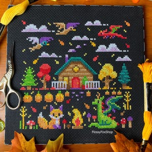May include: Cross-stitch art on black fabric featuring a whimsical autumn scene. A cabin, pumpkins, trees, and a fox are depicted. Dragons and clouds fill the sky. The text "FlossyFoxShop" is visible. Yellow thread and scissors are in the frame.