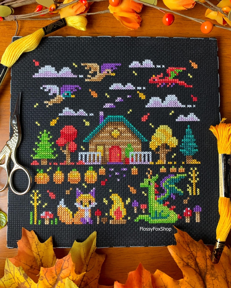 May include: Cross-stitch art on black fabric featuring a whimsical autumn scene. A cabin, pumpkins, trees, and a fox are depicted. Dragons and clouds fill the sky. The text "FlossyFoxShop" is visible. Yellow thread and scissors are in the frame.