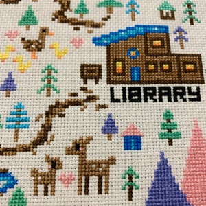 Puede incluir: Bordado de punto de cruz que representa una escena de invierno caprichosa con una caba&ntilde;a marr&oacute;n etiquetada como "LIBRARY", un lago azul y una variedad de &aacute;rboles y animales.