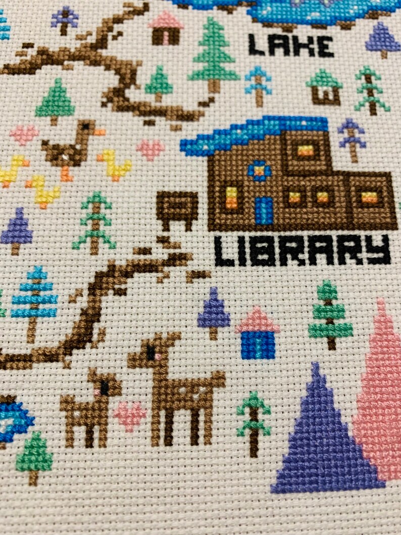 Puede incluir: Bordado de punto de cruz que representa una escena de invierno caprichosa con una caba&ntilde;a marr&oacute;n etiquetada como "LIBRARY", un lago azul y una variedad de &aacute;rboles y animales.