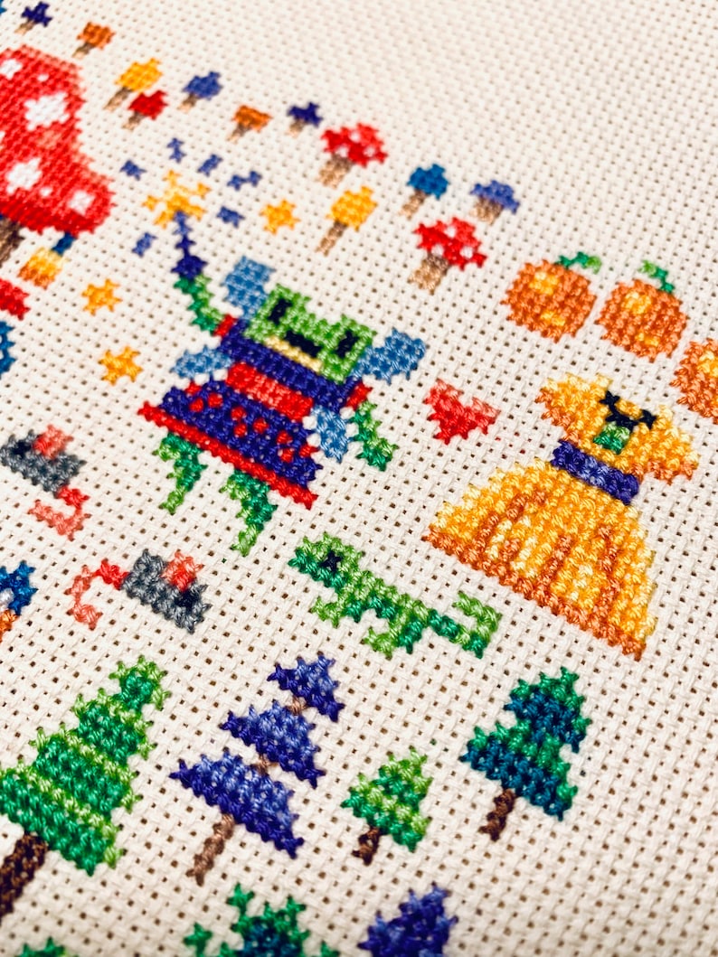 May include: Cross-stitch embroidery featuring a green and blue fairy with a red dress, a yellow dog with a blue collar, and pumpkins. The background includes trees, mushrooms, and stars.