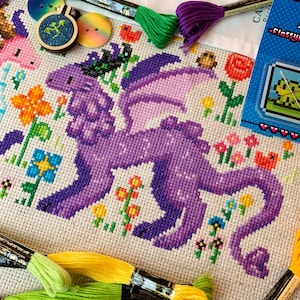 May include: A completed cross-stitch project featuring a purple dragon with lavender wings, surrounded by colorful flowers. Embroidery floss in green, yellow, and purple are visible, along with buttons and a small framed design. A pixelated frog design is also present.