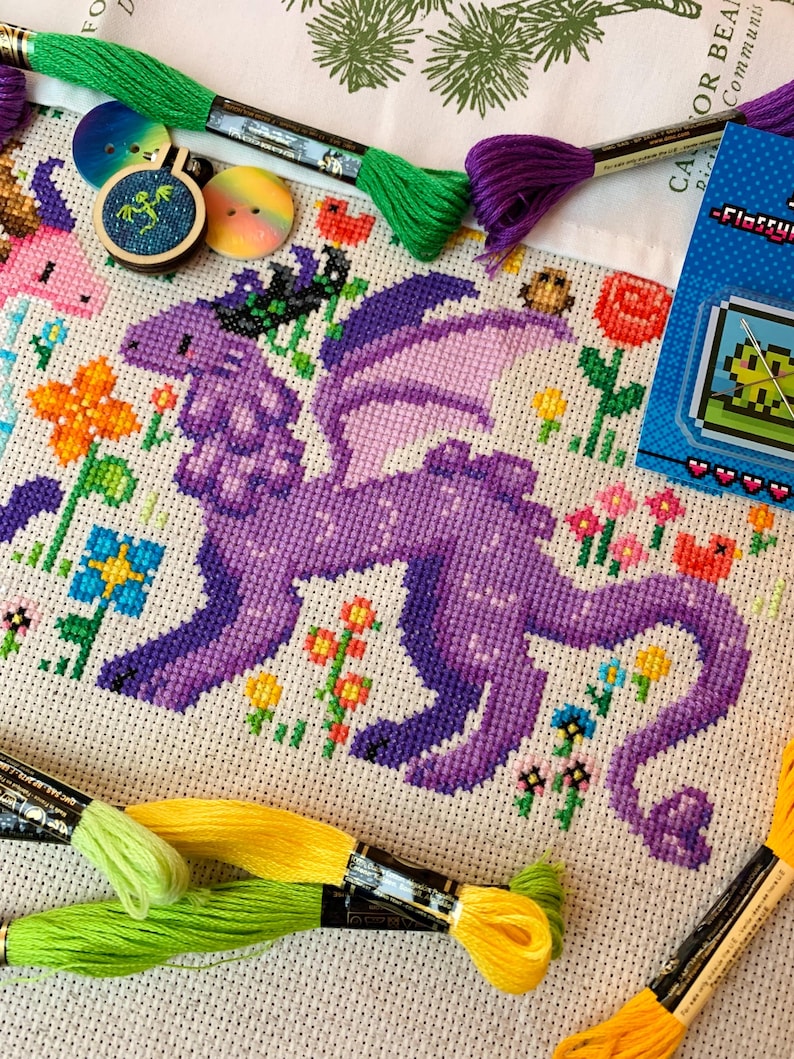 May include: A completed cross-stitch project featuring a purple dragon with lavender wings, surrounded by colorful flowers. Embroidery floss in green, yellow, and purple are visible, along with buttons and a small framed design. A pixelated frog design is also present.