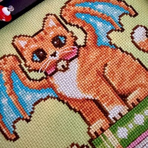 May include: Cross-stitch art featuring a cartoon cat with orange fur, blue wings, and a green and pink striped base. The cat has large blue eyes and a pink tongue. The background is a light green fabric, and the piece is framed in black.