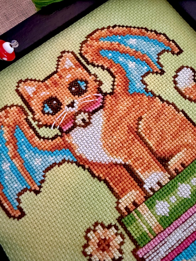 May include: Cross-stitch art featuring a cartoon cat with orange fur, blue wings, and a green and pink striped base. The cat has large blue eyes and a pink tongue. The background is a light green fabric, and the piece is framed in black.