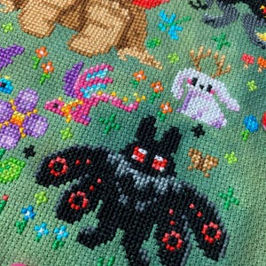 May include: Cross-stitch embroidery featuring a black bat with red eyes, a pink dragon, a white bunny, and a brown bear wearing a red scarf. The background is green with flowers and other small details.