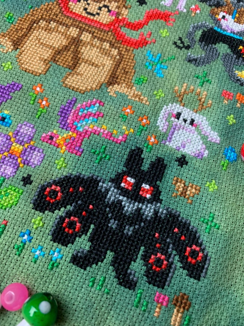 May include: Cross-stitch embroidery featuring a black bat with red eyes, a pink dragon, a white bunny, and a brown bear wearing a red scarf. The background is green with flowers and other small details.