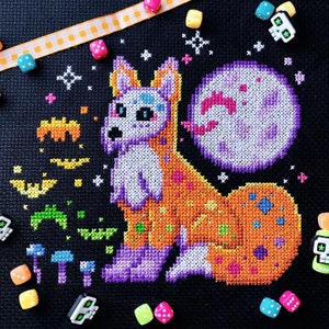 May include: Cross-stitch pattern of a purple and orange fox wearing a costume with a skull and crossbones design. The fox is surrounded by stars and a purple moon.