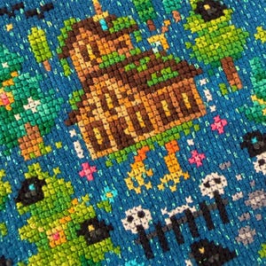 May include: A cross-stitch embroidery on a vibrant blue fabric featuring a brown wooden house surrounded by green trees. The design includes small white skulls, black bats, and colourful star-like accents scattered across the blue background.
