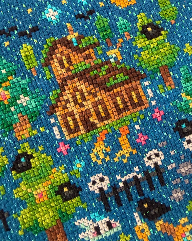 May include: A cross-stitch embroidery on a vibrant blue fabric featuring a brown wooden house surrounded by green trees. The design includes small white skulls, black bats, and colourful star-like accents scattered across the blue background.