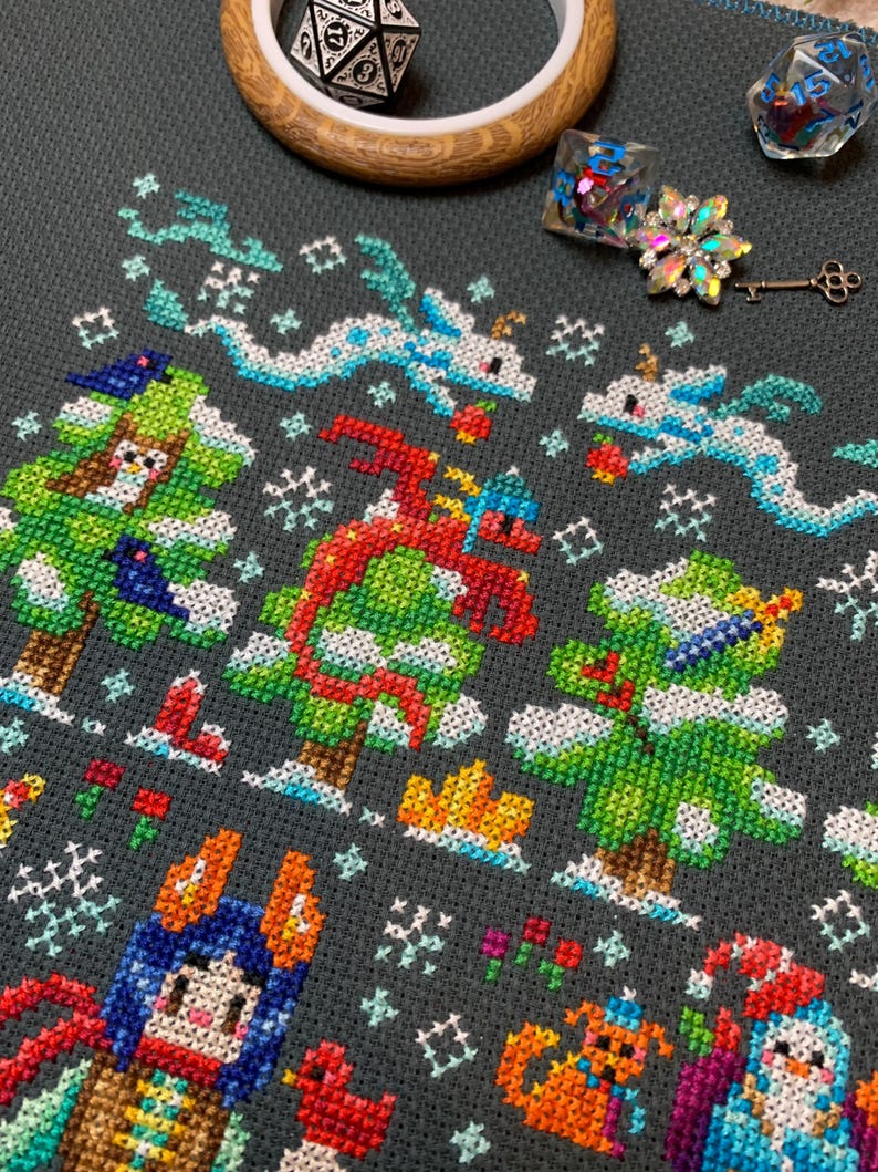 May include: A cross-stitch pattern featuring a winter wonderland scene with a red dragon, a green tree, a blue and orange character, and a white snowman. The pattern is on a dark gray fabric with white snowflakes.