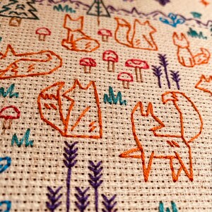 May include: Embroidered design on a beige fabric featuring multiple orange foxes, green trees, purple flowers, and red mushrooms.