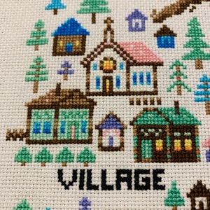 Puede incluir: Bordado de punto de cruz que representa una escena de pueblo con casas, &aacute;rboles y la palabra "VILLAGE" en letras negras.