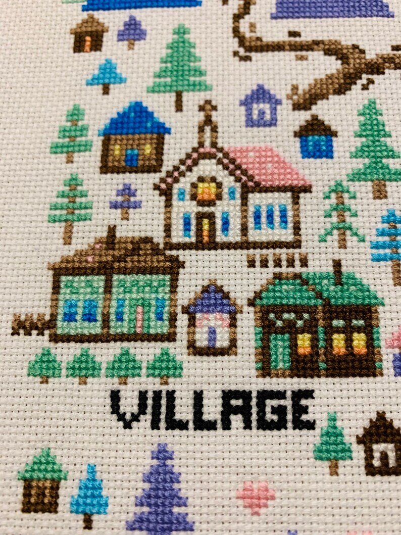Puede incluir: Bordado de punto de cruz que representa una escena de pueblo con casas, &aacute;rboles y la palabra "VILLAGE" en letras negras.