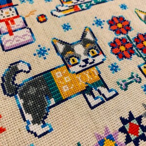 Peut inclure: Motif de point de croix repr&eacute;sentant deux chats portant des pulls. Le chat de gauche est gris et blanc avec un pull vert, jaune et bleu. Le chat de droite est orange et blanc avec un pull rouge et vert. L'arri&egrave;re-plan est un tissu de couleur cr&egrave;me avec des flocons de neige et des fleurs.
