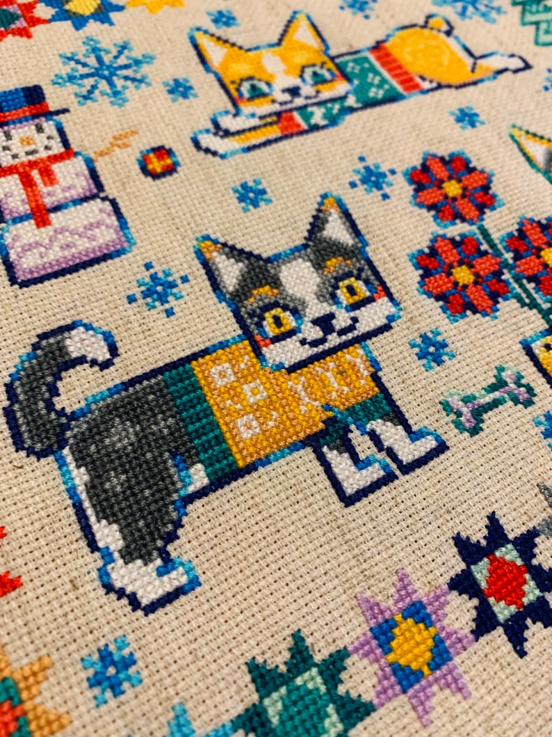 Peut inclure: Motif de point de croix repr&eacute;sentant deux chats portant des pulls. Le chat de gauche est gris et blanc avec un pull vert, jaune et bleu. Le chat de droite est orange et blanc avec un pull rouge et vert. L'arri&egrave;re-plan est un tissu de couleur cr&egrave;me avec des flocons de neige et des fleurs.