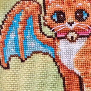 May include: Cross-stitch of a cartoon cat with orange fur, blue wings, and large eyes. The cat has white whiskers and pink cheeks. The background is a light green color.