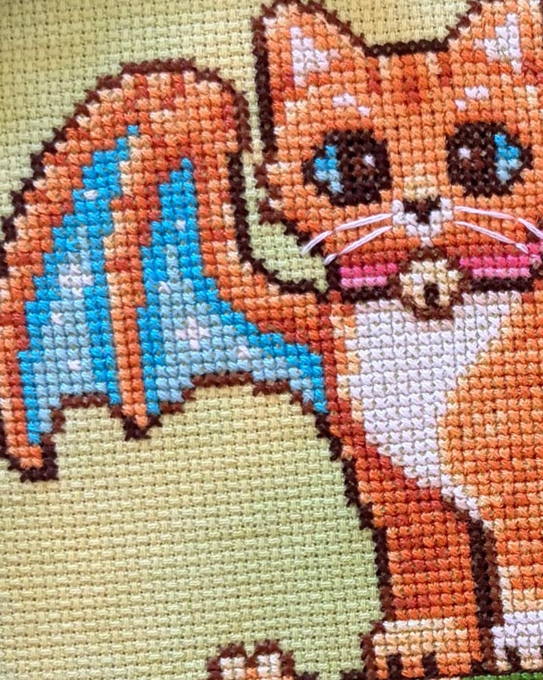 May include: Cross-stitch of a cartoon cat with orange fur, blue wings, and large eyes. The cat has white whiskers and pink cheeks. The background is a light green color.