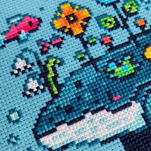 Puede incluir: Primer plano de un dise&ntilde;o de punto de cruz sobre tela azul claro. El dise&ntilde;o presenta una ballena con flores y peces pixelados de colores. La ballena es principalmente azul y blanca, con acentos de rosa, naranja y verde. Las flores y los peces tambi&eacute;n son de colores brillantes.