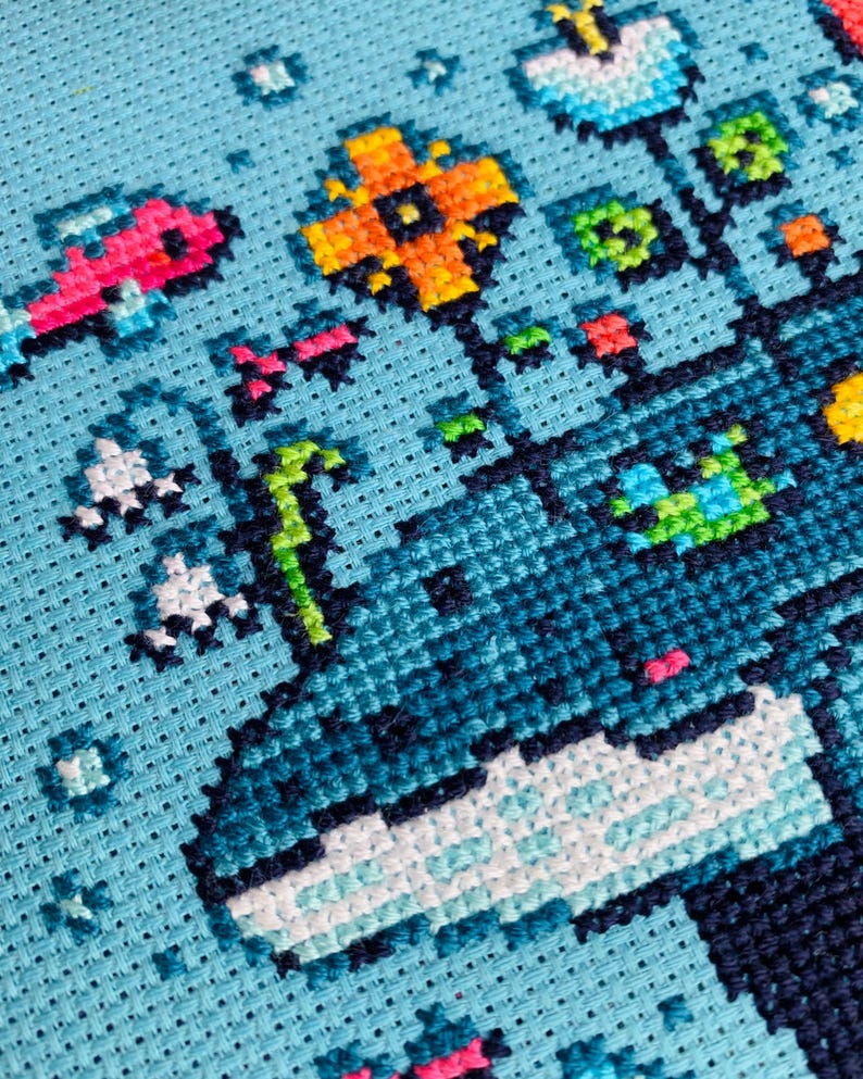 Puede incluir: Primer plano de un dise&ntilde;o de punto de cruz sobre tela azul claro. El dise&ntilde;o presenta una ballena con flores y peces pixelados de colores. La ballena es principalmente azul y blanca, con acentos de rosa, naranja y verde. Las flores y los peces tambi&eacute;n son de colores brillantes.