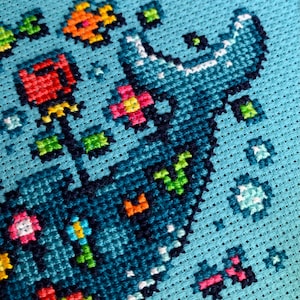 Puede incluir: Primer plano de un dise&ntilde;o de punto de cruz sobre tela azul claro. El dise&ntilde;o presenta una forma azul grande y estilizada con acentos blancos, rodeada de elementos florales y geom&eacute;tricos coloridos en rojo, naranja, verde y rosa. Las puntadas son claramente visibles.